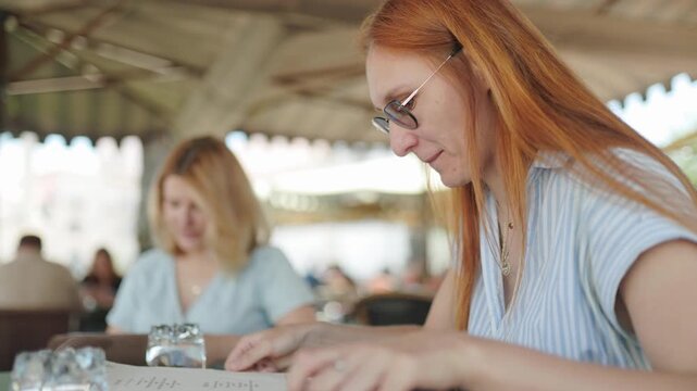 Two women look at menu while sitting at cafe table in sunny outdoor area