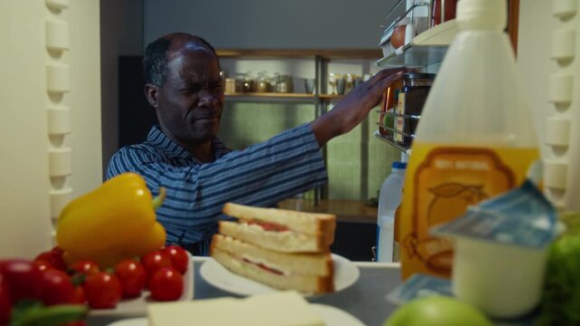 Senior African American man opening refrigerator, looking into camera with guilty expression and taking plate with sandwich. Ideal for late-night snacking or diet campaigns