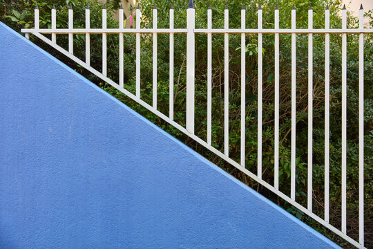 Diagonal ansteigende blaue Putzwand neben wei&szlig;en Metallzaun mit Speerspitzen vor Hecke.