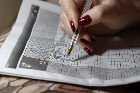 A person with red glittery manicured nails solves a Japanese nonogram logic puzzle using a ballpoint pen to represent deep concentration, cognitive development and mindful leisure