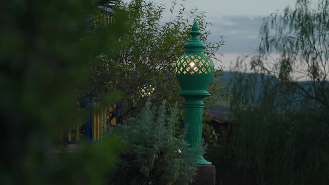 Ornate green metal garden lamp with pierced lattice glowing warmly among plants at dusk