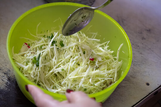 A hand holding a metal spoon stirs fresh shredded cabbage salad in a green bowl, reflecting healthy homemade meal preparation in a cozy kitchen setting for a nutritious lifestyle