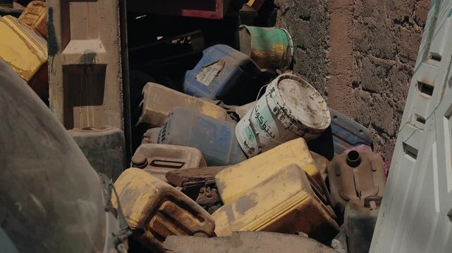 Weathered plastic jerrycans and paint buckets piled in a narrow gap by a mud wall