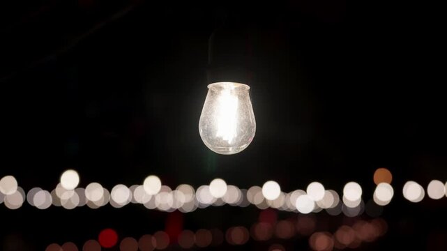 One lit festoon bulb hanging in the dark above a blurred row of white lights