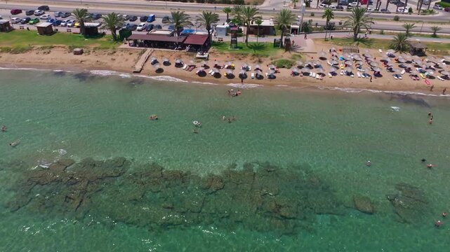 Aerial flyover directly above a row of beach umbrellas and sunbathers at Long Beach, Iskele, North Cyprus, with clear turquoise water, visible reef patches, and the coastal road alongside.