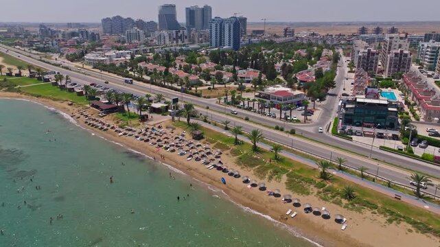 Aerial drone reveal sweeping along Long Beach in Iskele, North Cyprus, showing rows of beach club sunbeds and the coastal road, with the developing high-rise skyline in the distance.