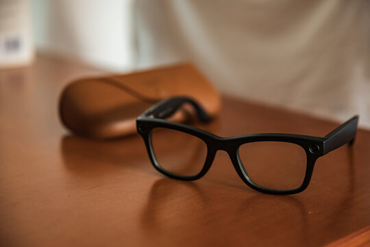 Gafas inteligentes de montura negra con c&aacute;mara integrada y estuche de cuero sobre mesa de madera