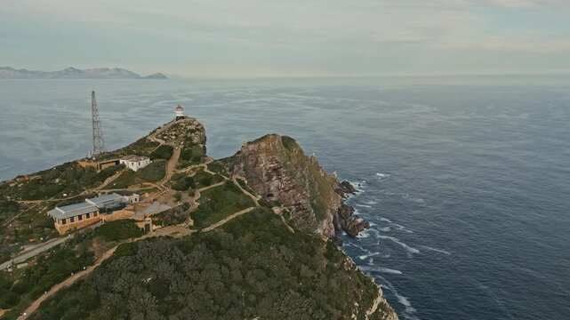 Drone push in reveals Cape Point Lighthouse above rugged cliffs and Atlantic Ocean in Cape Town, South Africa, with winding paths, rocky coastline, green fynbos and distant mountains beyond