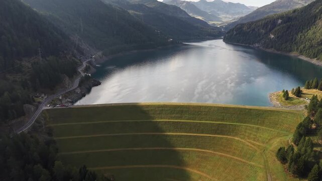 Aerial view of the Marmorera Dam, a large curved embankment dam holding back the Marmorera reservoir, surrounded by lush alpine forests and steep mountain slopes in Surses, Grisons, Switzerland.