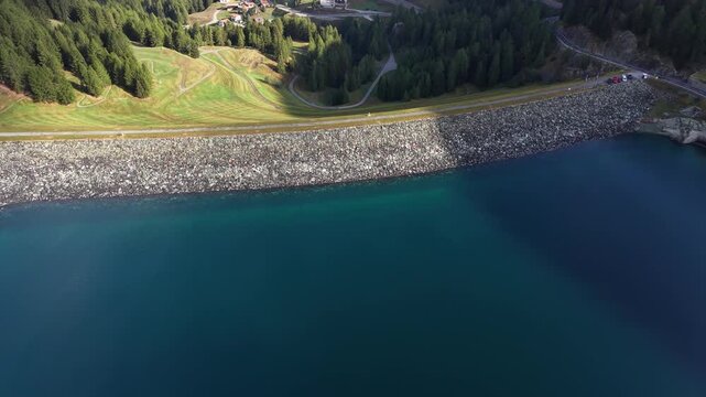 Aerial view of Marmorera Dam with its stone embankment and deep blue reservoir water surrounded by coniferous forests and green alpine slopes in Surses, Grisons, Switzerland.