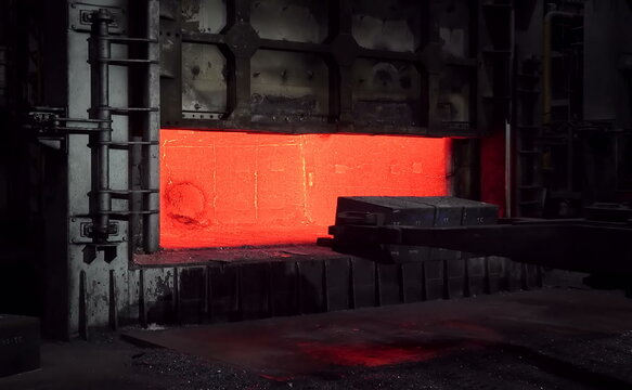 A large open furnace reveals a glowing red-hot interior, with thick metal walls and a mechanical arm positioned nearby, emphasizing high-temperature metal treatment and precision forging operations. 