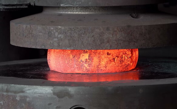 Industrial hydraulic press applying heavy pressure to shape a glowing hot metal block placed on the lower die within a heavy manufacturing facility workshop environment
