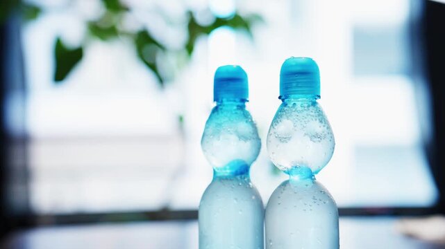 Two blue capped plastic bottles side by side with condensation, blurred green plant behind