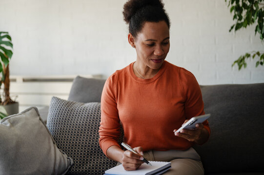Woman uses a smartphone while sitting at home with a notebook, representing mobile banking, budgeting and managing personal finances online