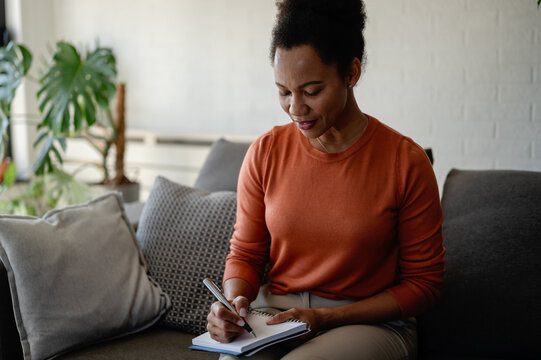 Professional woman writes notes while sitting on a sofa at home, working on plans, ideas and tasks in a relaxed remote work environment