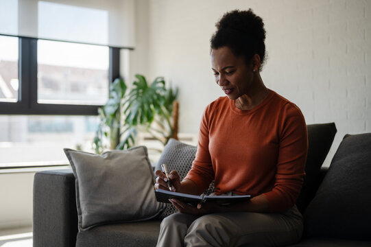 Woman writes in a notebook while relaxing at home, reflecting on ideas, setting goals and practicing journaling for personal development
