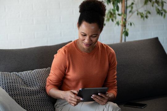 Relaxed smiling woman uses a tablet while sitting comfortably on a sofa at home, enjoying digital entertainment and leisure time