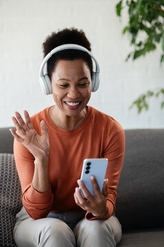 Happy woman wearing headphones waves while making a video call on her smartphone from home, enjoying friendly online communication