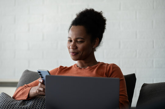 Relaxed woman uses a smartphone while working on a laptop from a sofa at home, combining mobile technology with remote work