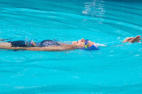 Senior woman wearing swim goggles and a cap, swimming backstroke in a bright blue pool, engaging in aquatic exercise for health and wellness
