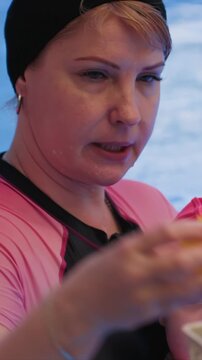 Child wearing red headwear stands near woman holding. Person in wetsuit manipulates small yellow object towards. Background shows aquatic environment suggesting proximity