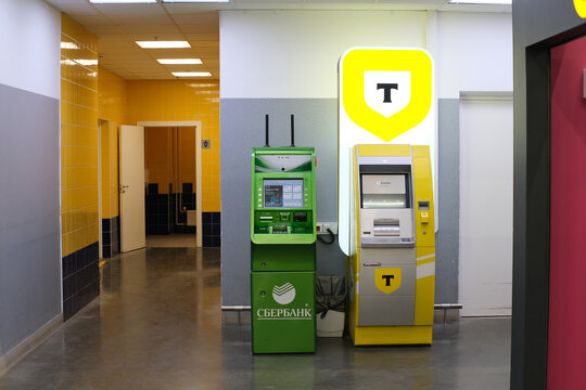 Two branded banking terminals in a colorful public building interior, featuring green and yellow ATMs, tiled walls, a hallway, and a white service door. Sber, Tbank