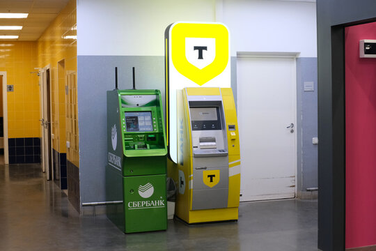 Two branded banking terminals in a colorful public building interior, featuring green and yellow ATMs, tiled walls, a hallway, and a white service door. Sber, Tbank