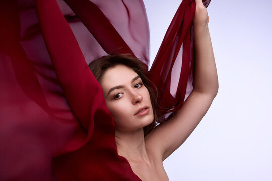 Young woman raising flowing burgundy fabric in graceful fashion beauty pose. Concept of fashion branding, fragrance ads and magazine editorial.