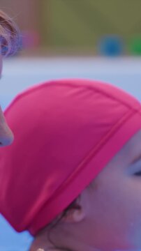 Woman and child in pink swim caps partially submerged. Focus remains tightly on side views of individuals. Outdoor shallow water area has green and blue objects