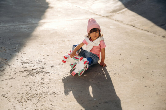 Young girl wearing pink beanie and pink shirt sits on concrete surface with roller skates, sunlight casting shadows around her in an outdoor skating area