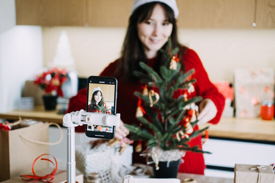 Phone on tripod records woman in Santa hat decorating small tree in kitchen. Social media christmas content, influencer decorating tree, vlogging holiday preparation smartphone