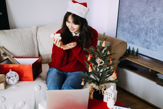 Young woman in Santa hat shows small toy ornament toward laptop while sitting on sofa near decorated Christmas tree. immigrant christmas abroad, diaspora holiday traditions, homesick during holidays