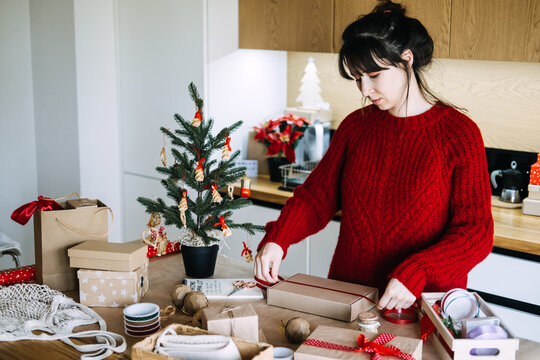 Woman pulls ribbon across kraft box on kitchen table beside decorated tree and wrapping supplies. Handmade versus mass produced, artisanal craft movement christmas, slow made gifts concept