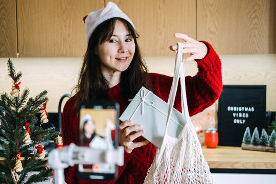 Woman in Santa hat shows gift box and mesh bag to phone camera near CHRISTMAS VIBES ONLY sign. Christmas gift guide video review, presenting gift ideas camera, holiday gift guide content creator