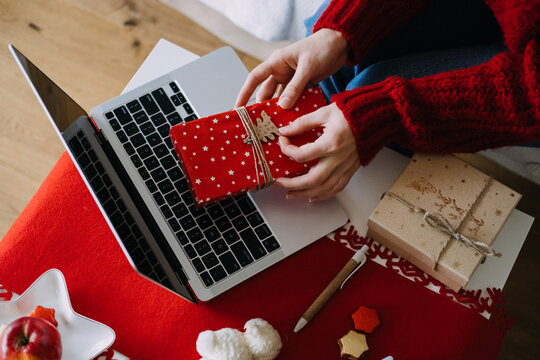Hands hold wrapped gift above laptop keyboard on table with parcels and pen. AI christmas gift recommendation, chatbot shopping assistant, personalized gift suggestions online