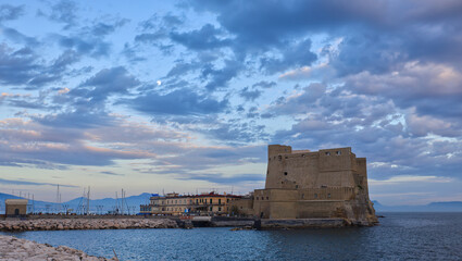serene castel dell'ovo standing tall in a coastal town at dusk, with boats and rocky shoreline in the foreground, brick and stone architecture, blue and gray tones, travel and history theme. © Stefano