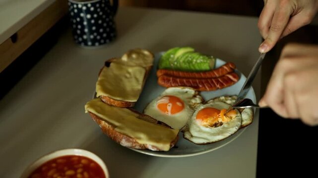 Close up view of a person using a knife and fork to eat a hearty breakfast plate featuring a fried egg sausage toast and avocado on a morning table.