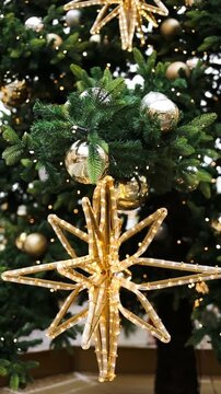Close up vertical shot of a beautiful golden shining star topper and elegant bauble hanging on a green Christmas tree branch with warm festive lights.