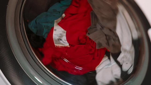 Close up of a modern white washing machine with a rotating glass drum filled with bright laundry spinning at home or laundromat.