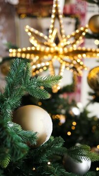 Close up vertical shot of a beautiful golden shining star topper and elegant bauble hanging on a green Christmas tree branch with warm festive lights.