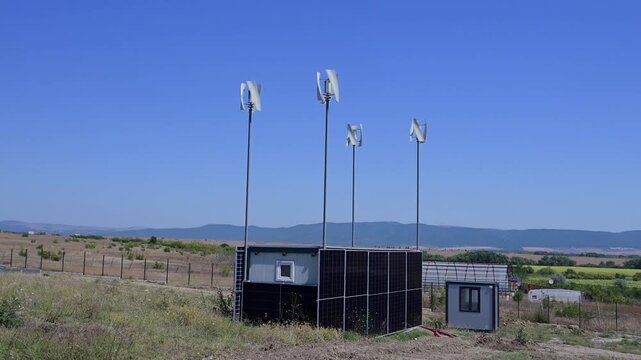 A small, off-grid container building clad entirely in solar panels sits in a rural field alongside four spinning vertical wind turbines under a bright, clear blue sky.