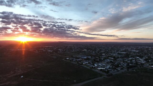 Sweeping aerial drone shot across Broken Hill at sunrise, with brilliant golden sunlight emerging beneath dramatic clouds over the surrounding outback landscape.