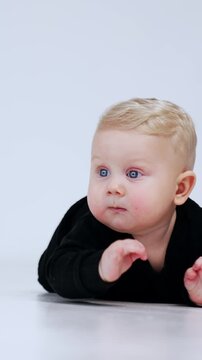 Adorable blond baby boy in black suit lies on the floor. Persistent kid crawls up to camera. White backdrop. Vertical video