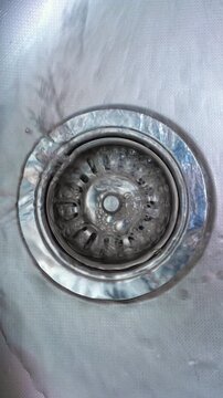 Water Flowing Into Stainless Steel Sink Drain. Close-up top view of clear water running across a metal basin and disappearing through the circular drain.
