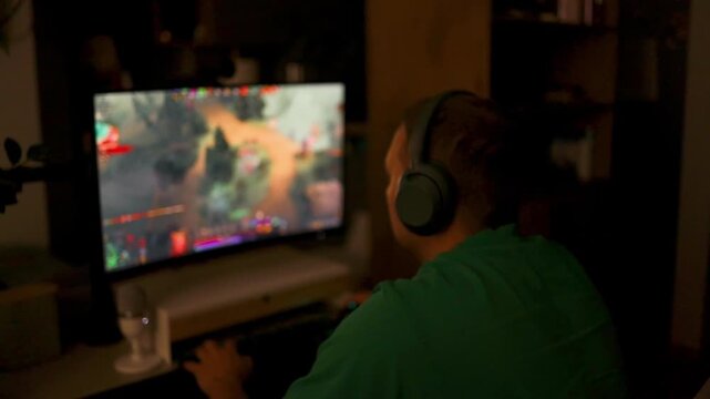 Over the shoulder view of a boy wearing headphones playing a colorful multiplayer strategy or shooter game on a monitor in a dark room.