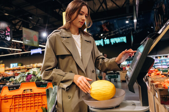 Young woman in beige trench coat weighing a melon on a digital scale inside a grocery store with fresh produce and shopping baskets visible in the background