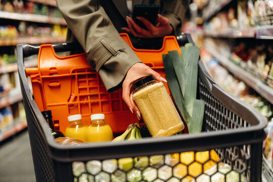 Female shopper in gray coat holding jar of spices while using smartphone in grocery cart filled with vegetables and beverages in supermarket aisle