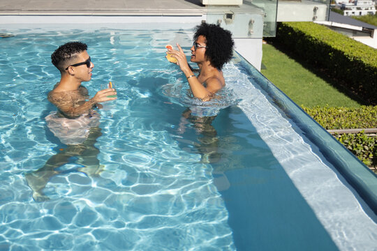 African American couple standing in rooftop pool in swimwear with sunglasses, glasses holding cups