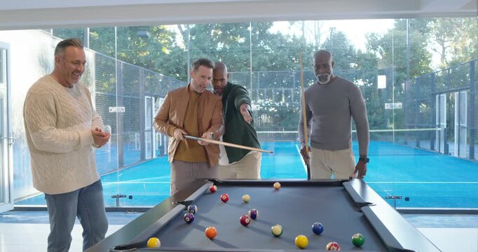 Diverse men coaching shooter, green shirt pointing with balls scattering on pool table by glass