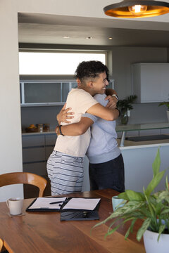 Male friends hugging near wooden dining table in modern kitchen with mug folder tablet potted plant